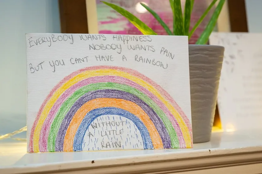 Hand-drawn rainbow card on shelf with handwritten message about happiness, pain, and hope, placed beside a potted plant and soft decorative lights indoors warmly cozy.