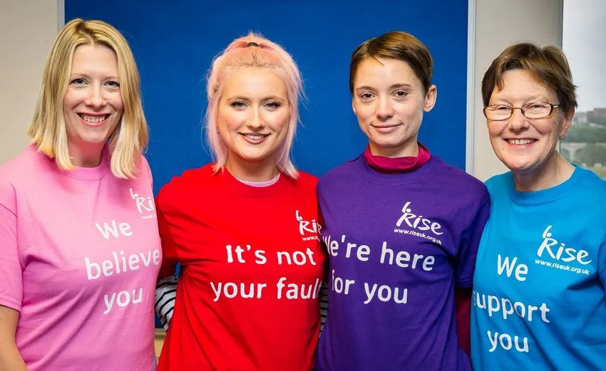 Four people stand side by side wearing colourful RISE T-shirts reading supportive messages, including “We believe you” and “We’re here for you,” against blue background.