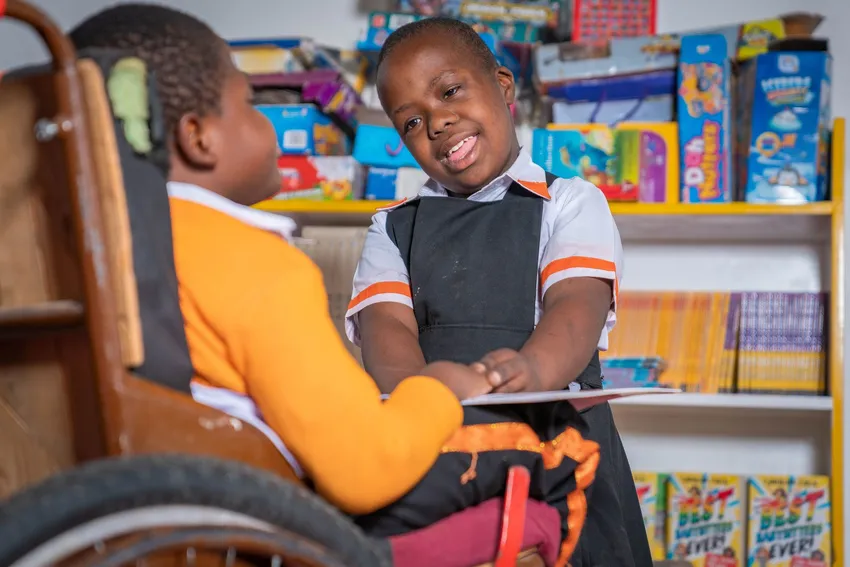 Girl standing up in front of a child in an orange jumper, sitting in a wheelchair.