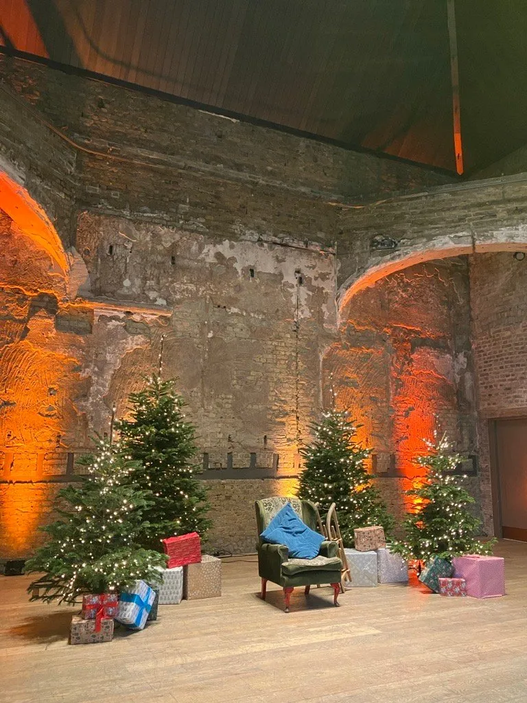 Decorated Christmas trees, wrapped presents and a green armchair set in BAC's historic Grand Hall for Community Christmas Day 2023, image credit Meg Khor