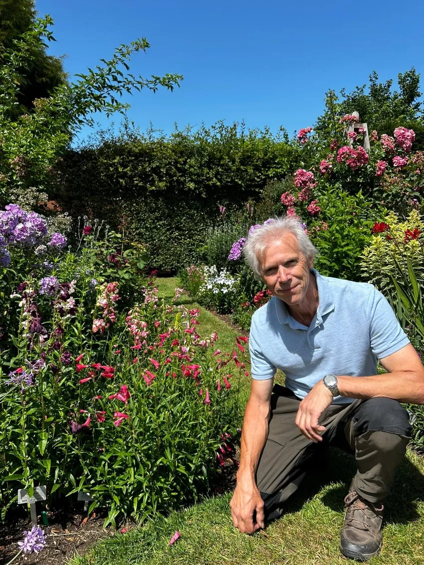 Richard kneels on a lawn in a colourful garden, tending to vibrant pink, purple, and red flowers which are part of his National Collection of Penstemon.