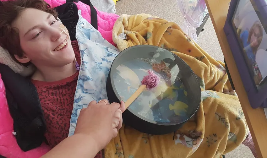 A young female wheelchair user is led watching a virtual Soundabout music session on an iPad. She's being helped to play an ocean drum on her lap.