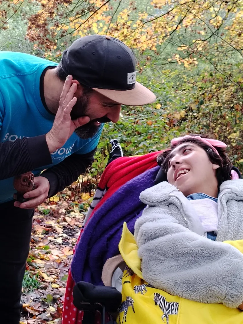 A young female wheelchair user is looking up and smiling at a male music practitioner who is leaning over and signing to her.