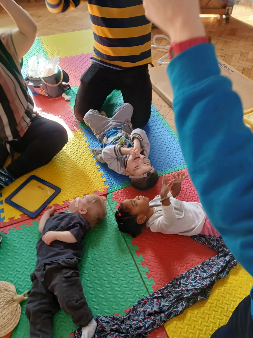 Three toddlers lie on colourful floor matts with adults holding kneeling around them holding their arms in the air.
