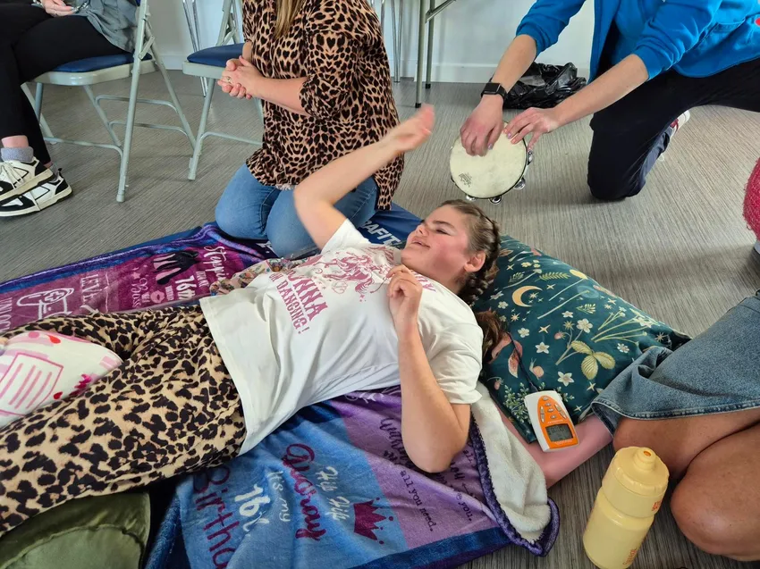 A young female Soundabout participant lies on a floor matt covered in blankets. She's smiling while others kneel around her playing instruments.