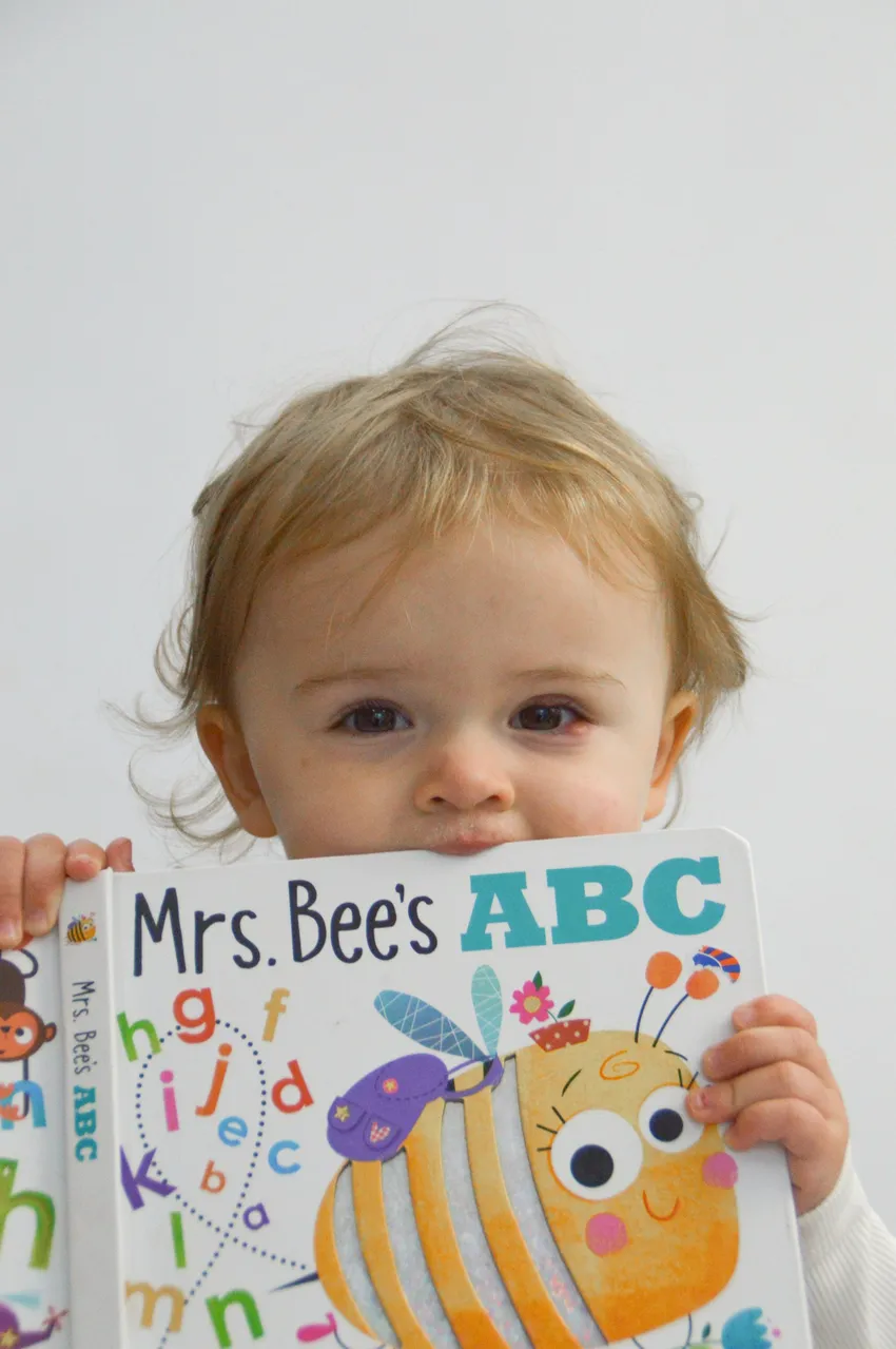 A little girl peeping over an ABC book with smiling eyes