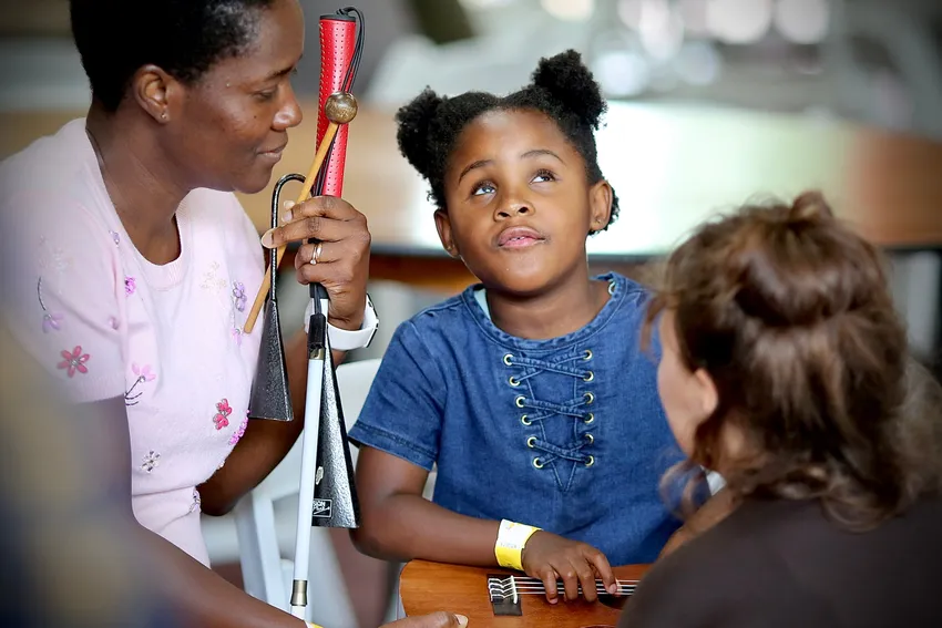 A visually impaired young person and their mother being taught how to strum a guitar. The mother is holding the young persons cane.
