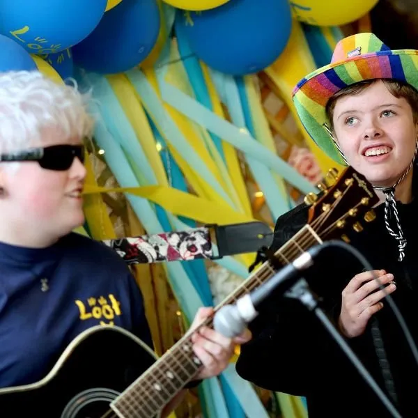 Two young adults performing on a stage, one is singing while the other plays the guitar.