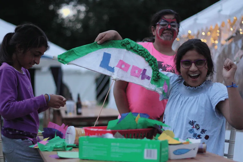Three children are gathered around a table covered with craft supplies, creating a sign that says "Look". Everyone is smiling and looks happy.