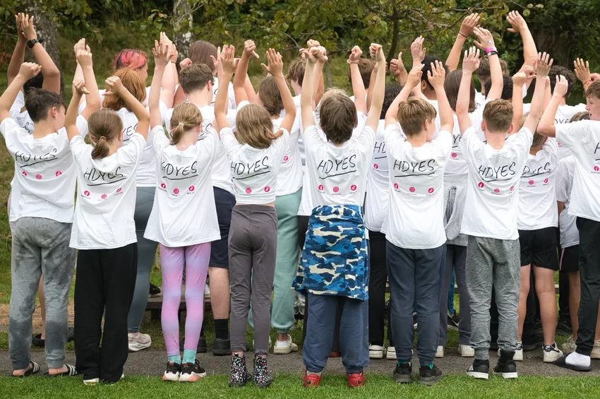 A group of children facing away from the camera with their arms in the air. They are all wearing t-shirts with the HDYES logo on the back.