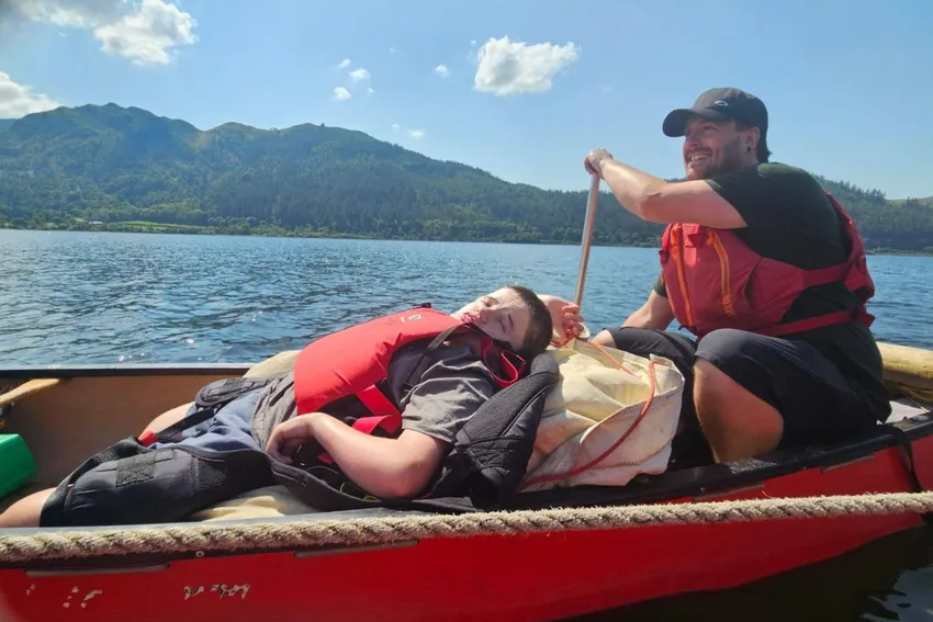 Young man resting on a reclined padded cushion in an open canoe on Bassenthwaite Lake with an instructor behind paddling