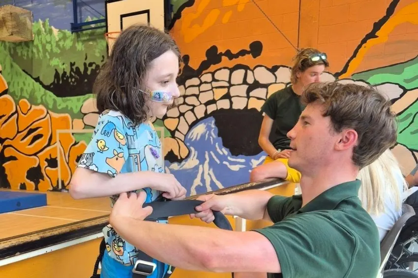 Young boy with a nose tube getting fitted for a harness by instructor Ben in an indoor sports hall