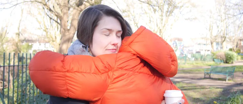 Two women are stood in a local park. They are hugging. The woman whose face is visible is smiling.