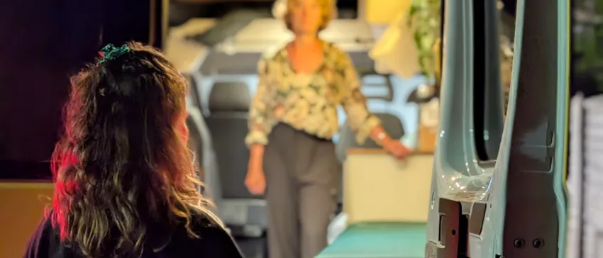 A woman stands behind One25's yellow outreach van, looking in through the open door. The interior is lit and a volunteer stands next to a bench seat, welcoming the woman in.