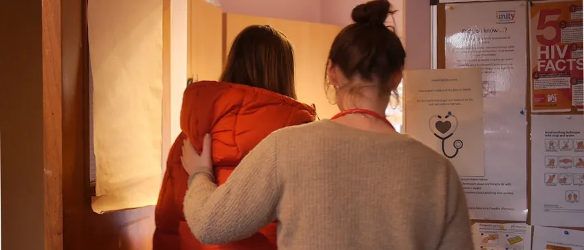 A woman in an orange coat walks into the GP surgery at One25. The GP, wearing a red stethoscope around her neck, stands beside the woman, with a hand on her shoulder.