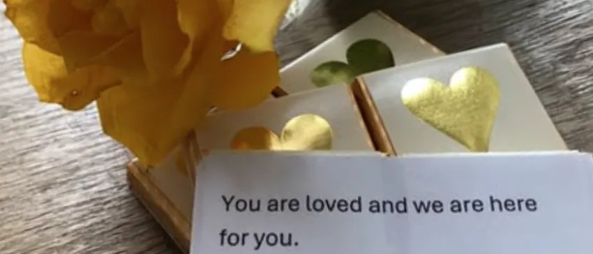A yellow rose is lying on a wooden table, next to a typed note that says, "You are loved and we are here for you."