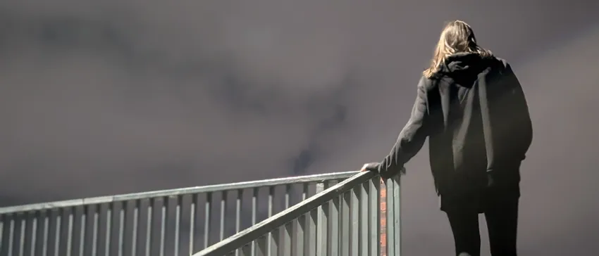 A woman wearing a black hooded sweatshirt stands at the top of a flight of stairs, leading to an overpass. The sky behind her is dark and cloudy.