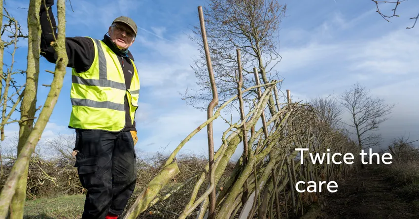 A man in a high‑visibility vest stands beside a freshly laid hedge made from interwoven branches. His hand rests on the hedge, and leafless trees and blue sky stretch across the background. The words “Twice the care” appear on the right side of the image.