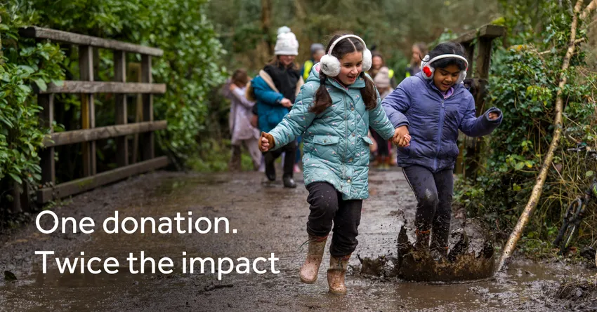 Two girls wearing coats and ear-warmers run on a muddy path, splashing in a large puddle. Their school friends walk behind them with trees and a wooden bridge framing the scene. The text “One donation. Twice the impact” appears on the left of the image.