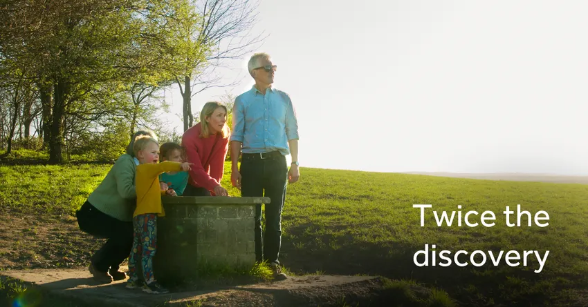 A family stands around a stone viewpoint on Wittenham Clumps, looking out toward a bright landscape. Trees frame the scene, and sunlight creates a warm glow. The text “Twice the discovery” appears on the right of the image.