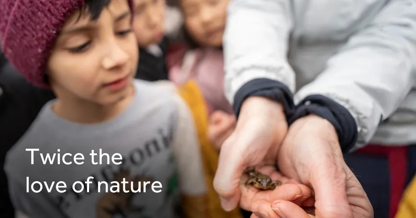 Hands hold a small frog, while several children stand close by observing. They are outdoors, with people dressed in warm clothing. The text “Twice the love of nature” appears on the left of the image.