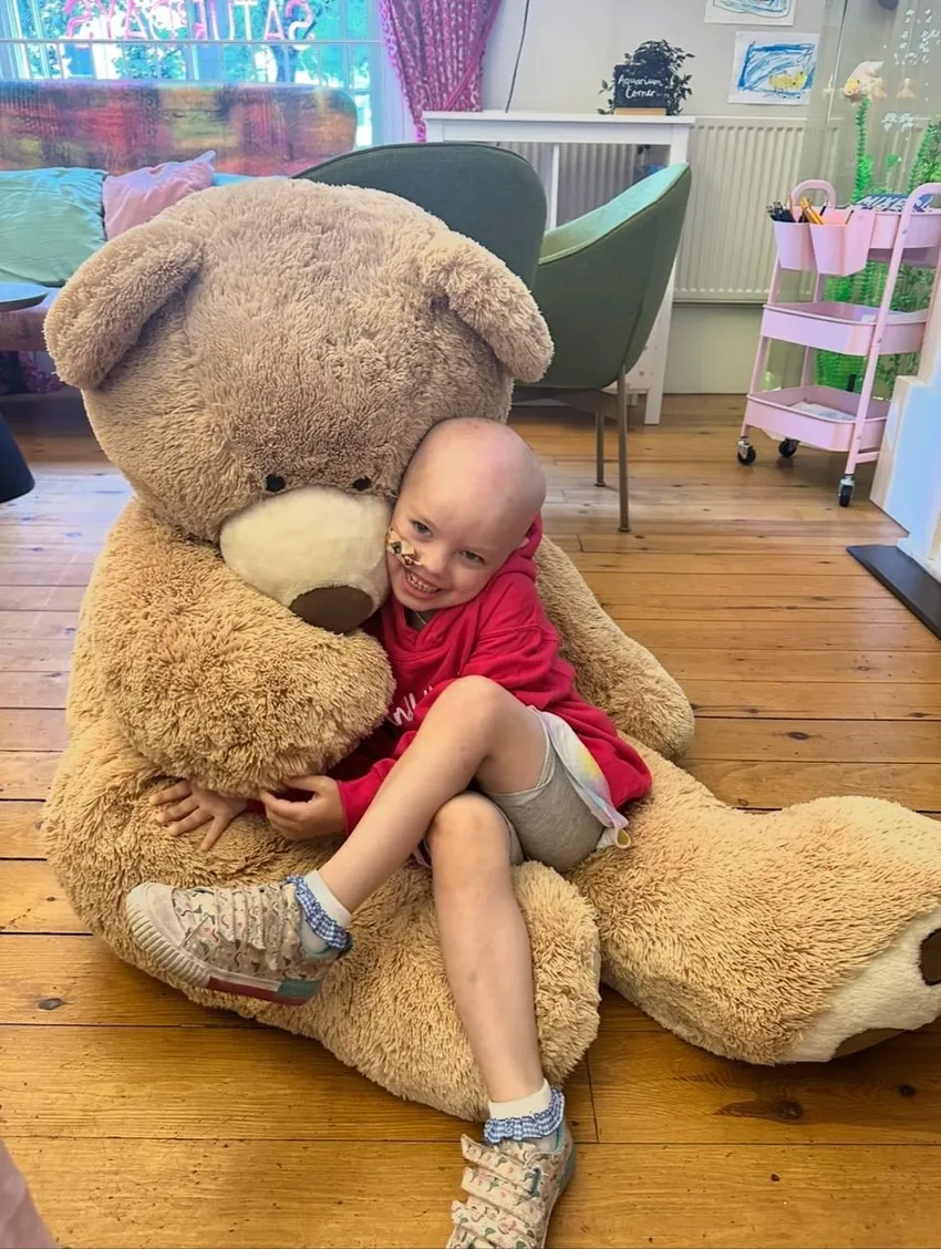 Young patient sitting in Candlelighters hoodie in family support centre. She is cuddling a huge teddy bear which is bigger than her.