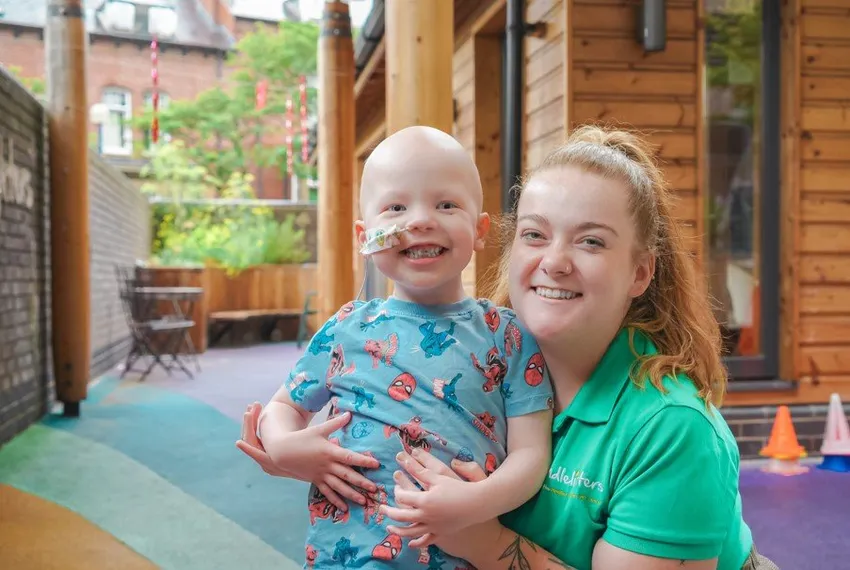 A young boy with a nasogastric tube, wearing a blue Spider-Man t-shirt, smiles broadly with the Candlelighters Youth Activity Coordinator in a garden just outside the ward