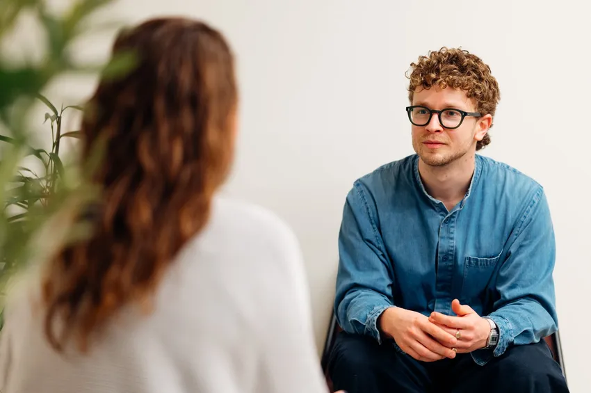 a relaxed man wearing glasses is sat facing a woman, listening attentively.