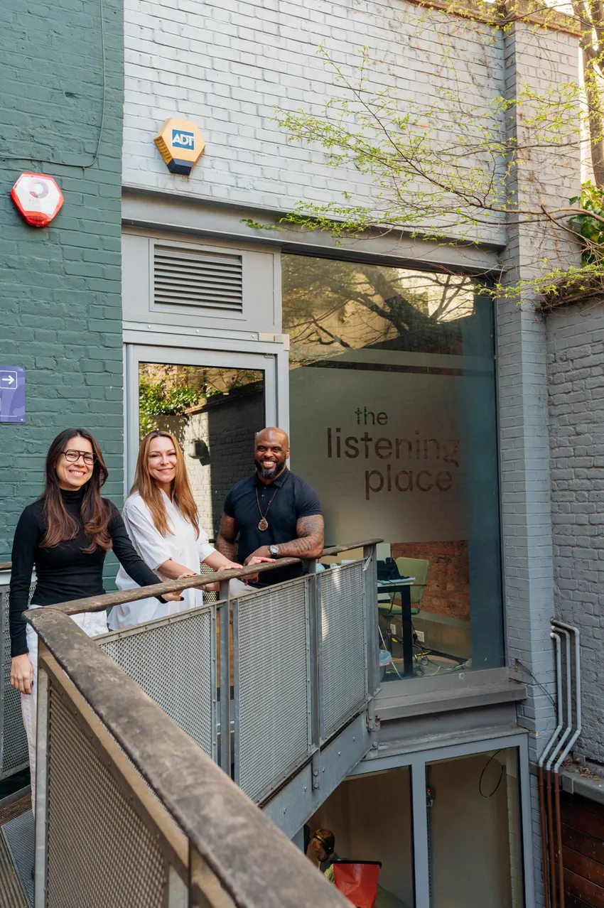 three people are standing on a metal balcony outside a building. Behind the group, there is a large glass window with the words "the listening place" visible inside. The three individuals are smiling and facing the camera.