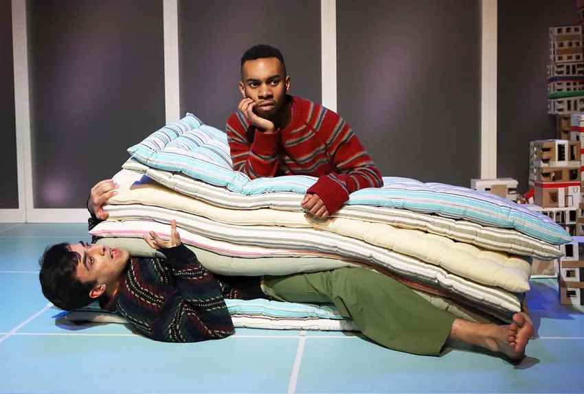 Two actors on a stage with a stack of mattresses: one man lies comically squished underneath them, while a Black actor in a red striped sweater rests his chin on the pile, looking down with concern.