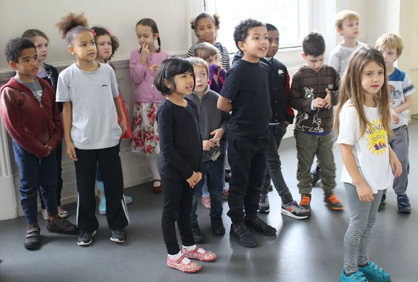 A group of diverse young children stand gathered together in a bright indoor room with pale walls. They are dressed in casual clothing, looking attentively off-camera in the same direction, appearing engaged in a group activity or instruction.