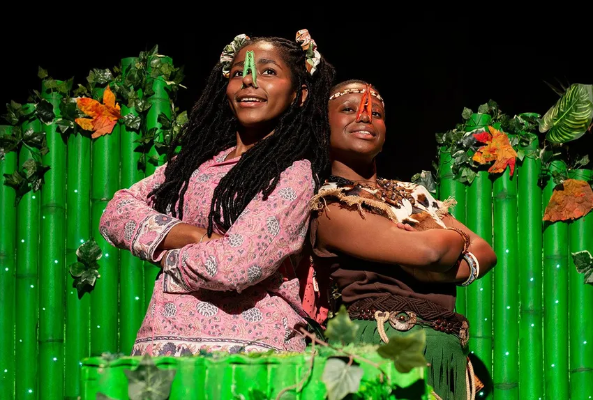 Two young Black actors stand back-to-back with arms crossed on a vibrant theater stage, each wearing a colorful clothespin on their nose. They are surrounded by bright green, bamboo-like scenic structures decorated with artificial leaves.