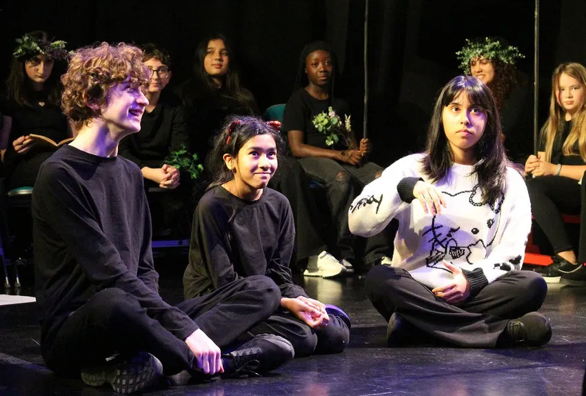A group of diverse young people are seated on a dark stage during a Youth Theatre sharing performance