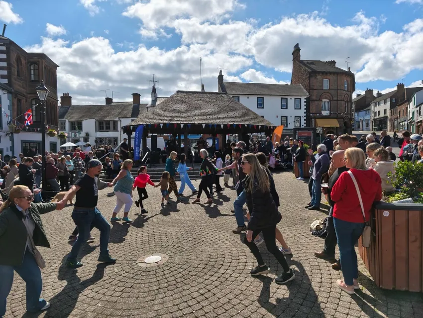 Children and adults dancing together in an outdoor ceilidh in a town square. Run by BlueJam Arts, in Penrith, on May Day 2025