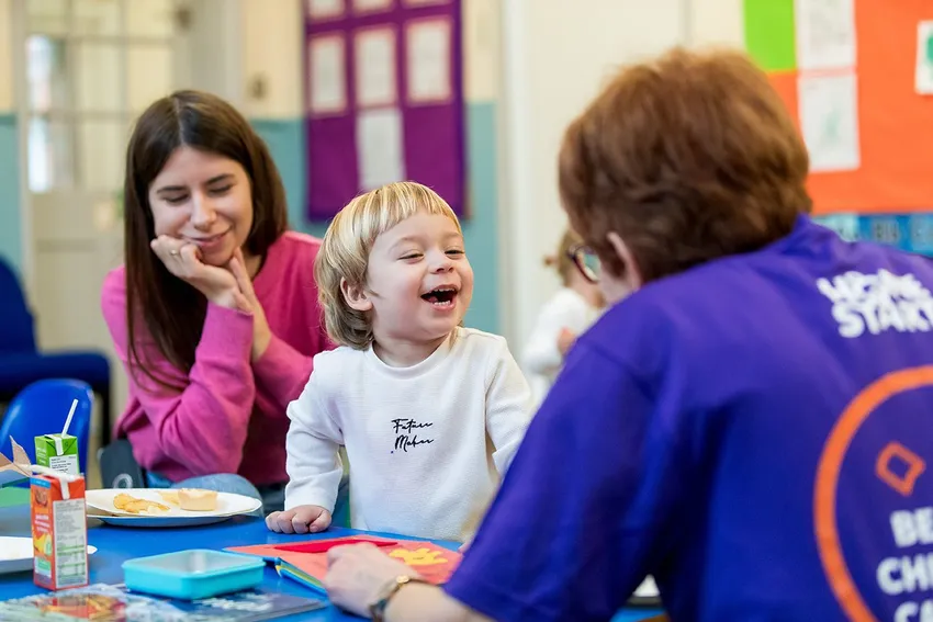 A Mum and young child smiling, enjoying some snacks and reading a book with a volunteer at one of Home-Start Banbury, Bicester & Chipping Norton's group sessions.