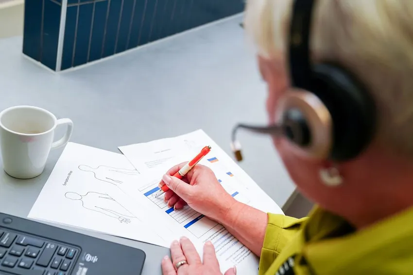 The back of Neuro Nurse Michaela's head with her headset on, taking notes on a symptoms tracker with an empty cup in front of her.