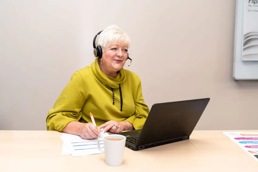Helpline Neuro Nurse Michaela, a lady with cropped white hair in a green roll neck top, is sitting at a desk with a cup of tea and laptop, speaking to someone on her headset as she guides them through some information.