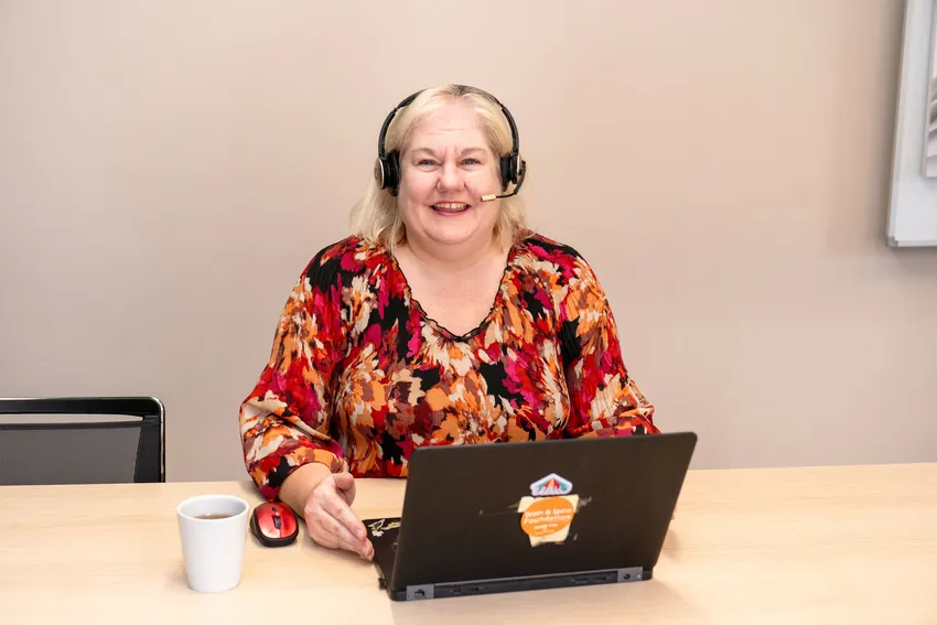Helpline Neuro Nurse Jo, a blond haired lady in a colourful top, is sitting at a desk with her headset on, cup of tea and laptop in front of her, ready to take calls from anyone affected by a neurological condition.