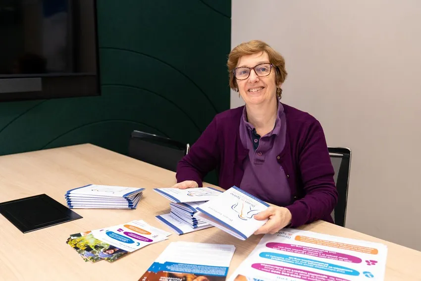Helpline Neuro Nurse Gail, with short cropped brown hair in a purple top and cardigan, is holding Brain and Spine Foundation information booklets in her hand with more booklets and leaflets on the table in front of her.