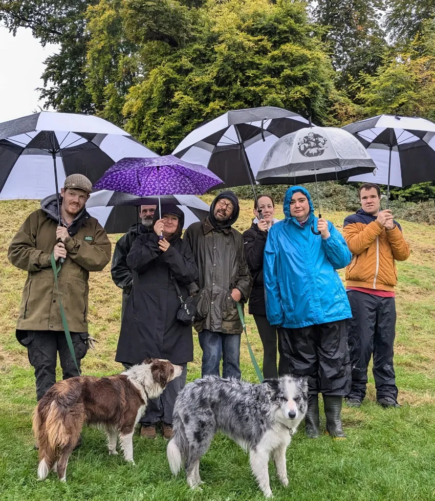 a group of smiling people under umbrellas in the countryside with two collie dogs in front of the group