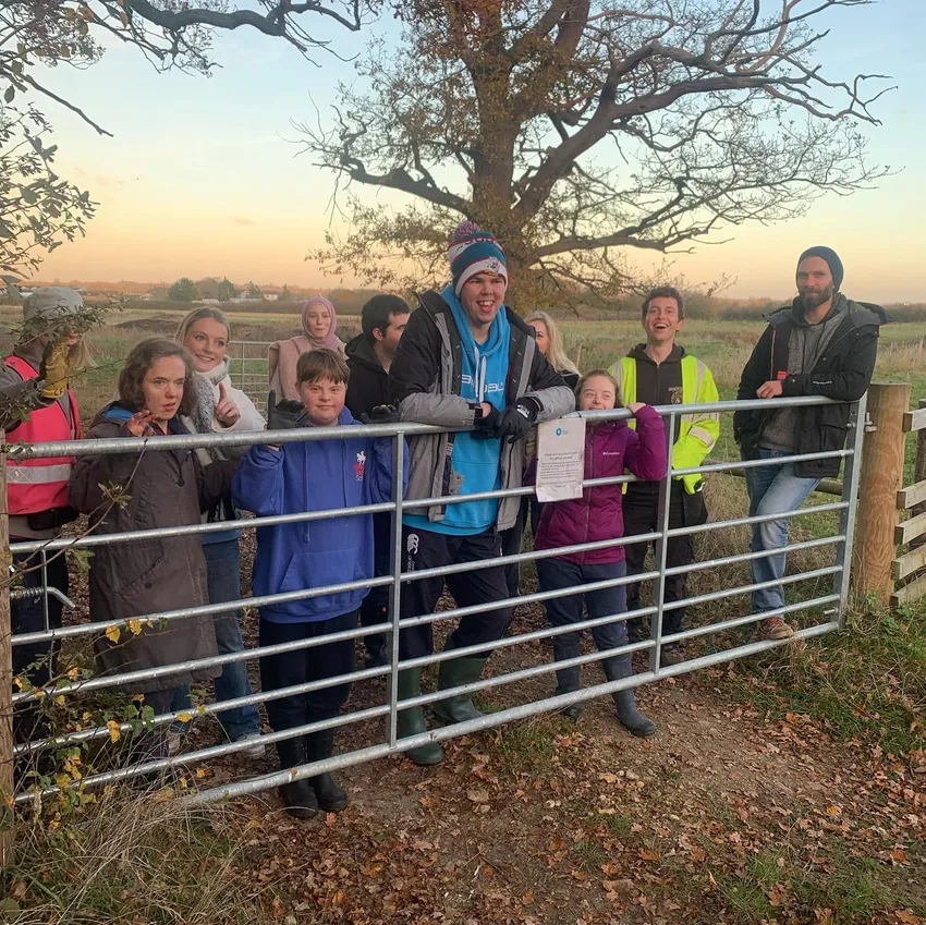 a mixed group of people standing peacefully behind a farm gate with a pink, dusky sky behind them
