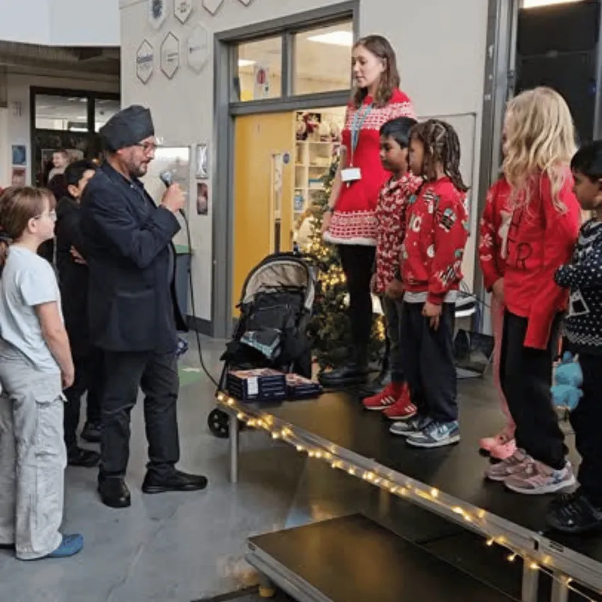 Children stand on a small, fairy‑lit stage in festive jumpers while an adult speaks into a microphone nearby.
