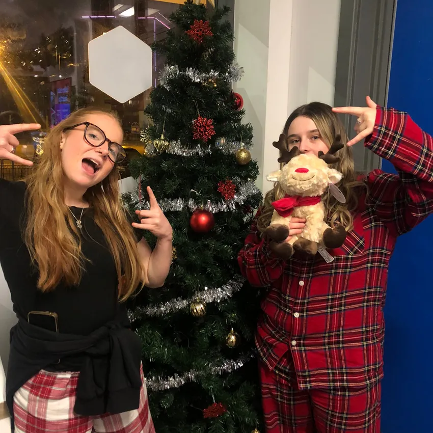 Two young people stand beside a decorated Christmas tree. One wears a black top with a jumper tied around the waist, while the other wears red tartan pyjamas and holds a soft toy reindeer. Both make playful hand gestures towards the camera.
