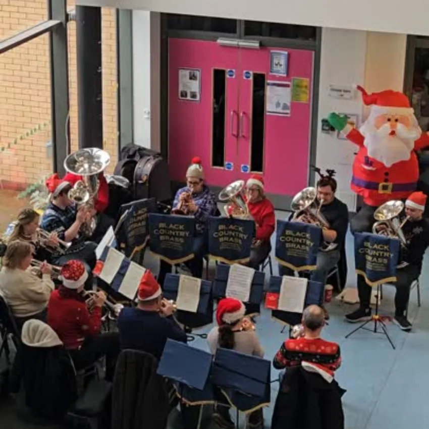A brass band wearing Christmas hats performs indoors at a Family Christmas event. Several people sit in the foreground watching the performance, also wearing festive clothing.