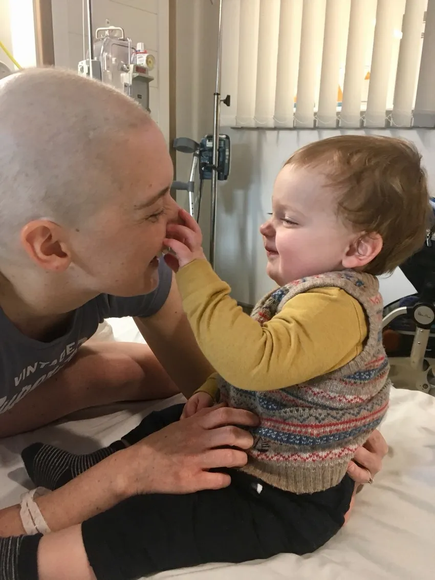 A mum with cancer is sitting next to her baby boy, who is propped next to her on a hospital bed. She has a bald head due to treatment. Her son has ginger hair and is wearing a cute jumper vest. He is grabbing her nose and she is laughing.