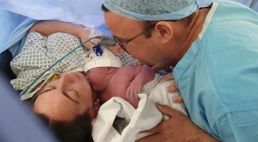 A mum is on the surgery table shortly after a c-section. We can just see her head, which has dark brown hair and lots of blue sheets. Her newborn baby is being held close to her face, by her husband, who is dressed in hospital scrubs.