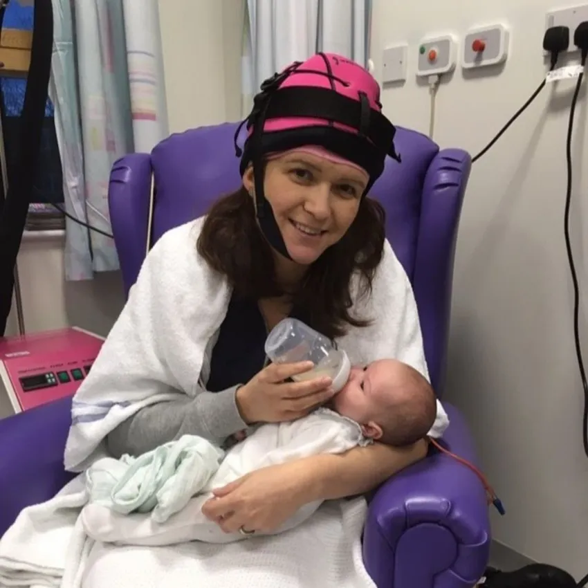A mum wearing a pink chemo cold cap and a hospital robe is sitting in a purple chair feeding her newborn with a bottle.