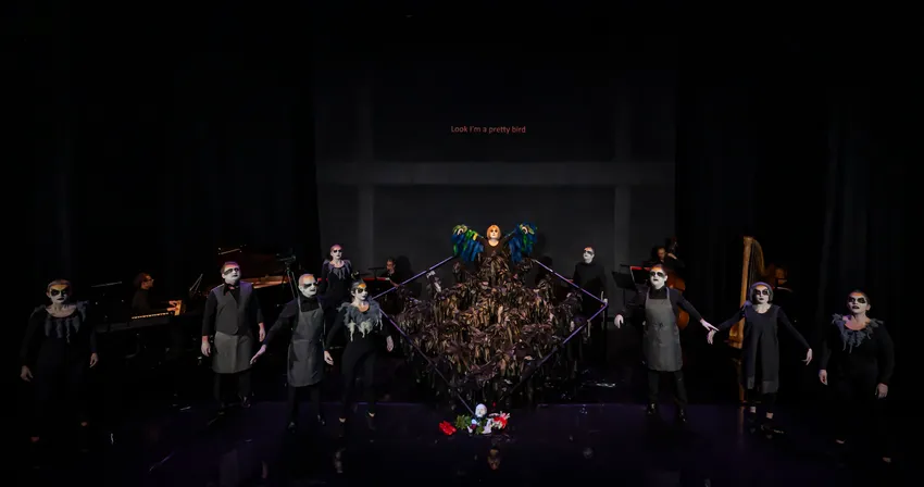 The image shows a black box theatre space, with the cast standing in a line across the front of the stage, and the pianist and band at the back.