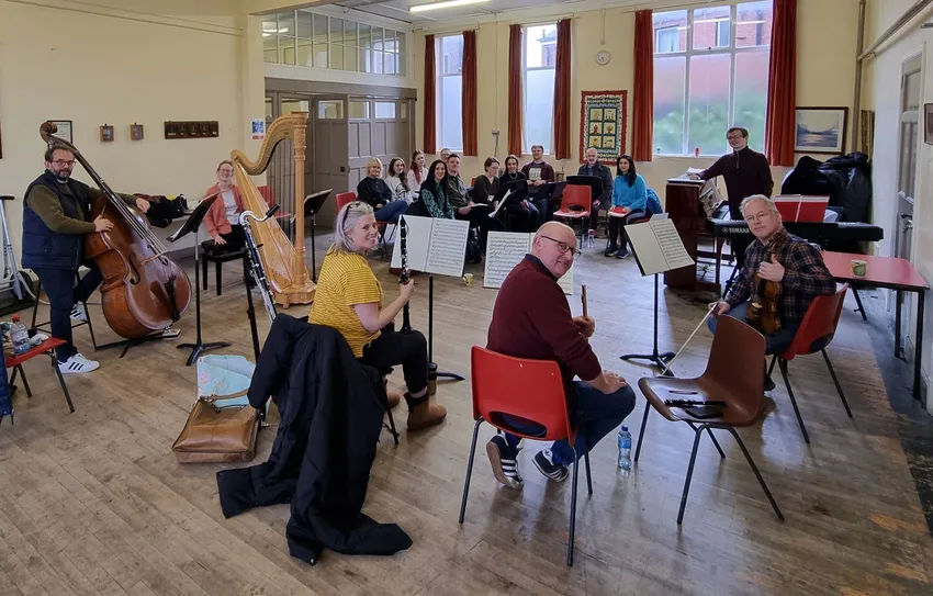 A pianist stands at a piano while a small orchestra and singers look at the camera, smiling. This was a rehearsal for a performance of a chamber opera by Philip Glass, musically directed by a pianist in training with NI Opera