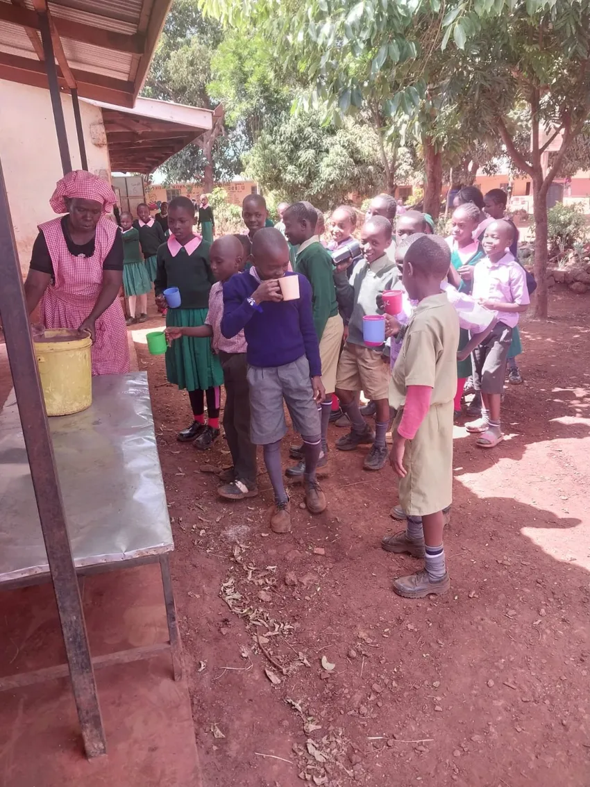 A group of around 20 Kenyan children in school uniform, queuing to have their plastic mugs filled with porridge, by a school cook in red apron and hat.